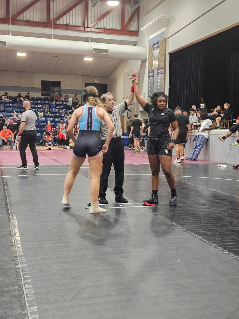 A wrestling official holds up Heather Butler's arm after she won a wrestling event against another wrestler. They stand in a crowded gym on top of wrestling mats.