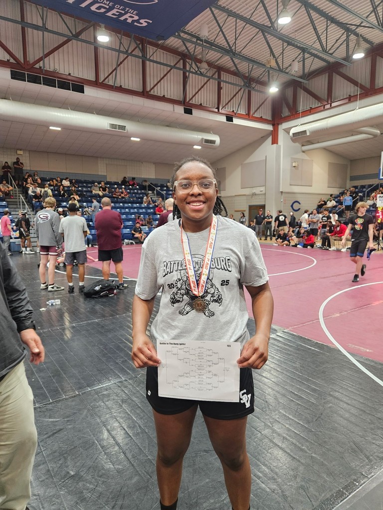Heather Butler smiles while holding a bracket and wearing a 1st place medal inside a gym with wrestling mats.