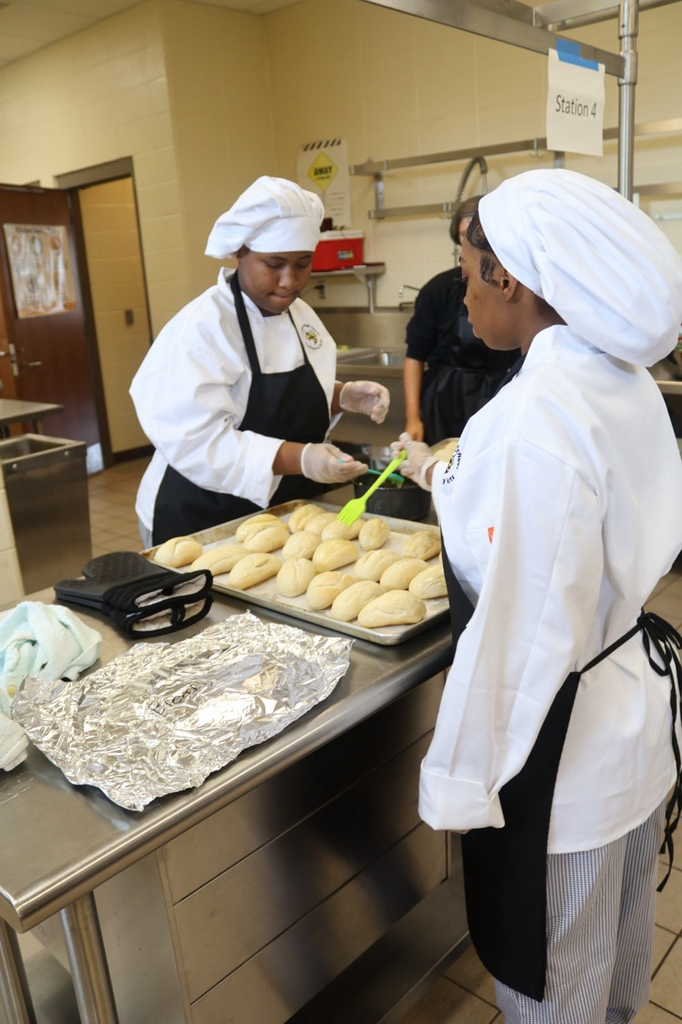 McAdort High School culinary students put butter on rolls while inside a kitchen.