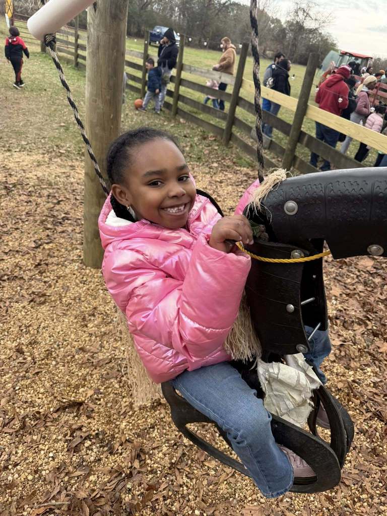 A student at a Christmas tree farm smiles while riding a tire swing decorated to look like a horse. 