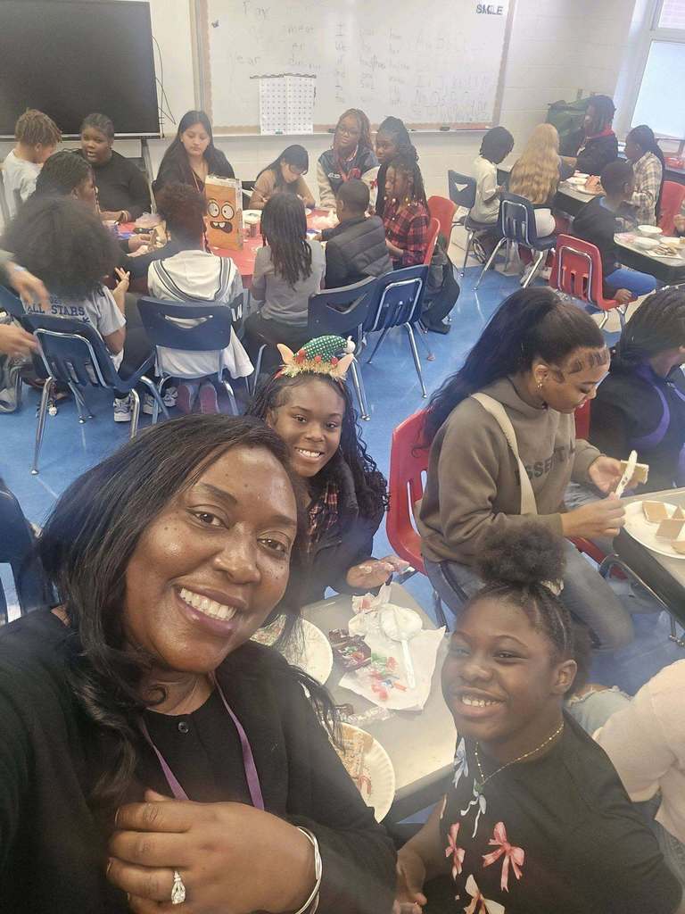 A woman and students smile for a photo while making gingerbread houses inside a school classroom.