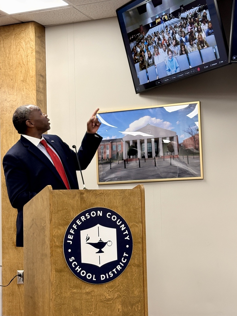 CCHS Principal Dr. Eugene Dallas points at a tv screen showing a video call with CCHS football players. He addresses the players from behind a podium in the Jefferson County Schools board room.