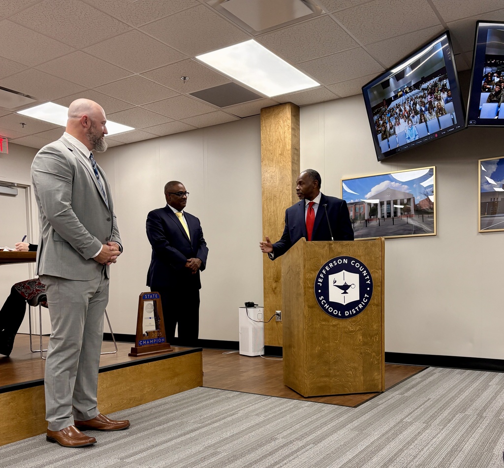 Clay-Chalkville High School Football Coach, Stuart Floyd,  CCHS Principal Dr. Eugene Dallas, and JEFCOED Superintendent Dr. Walter Gonsoulin stand together inside the Jefferson County Schools board room. Dr. Dallas speaks from behind a podium, and to the right of the photo, Clay-Chalkville's football players can be seen on a tv screen as they watch the meeting live.