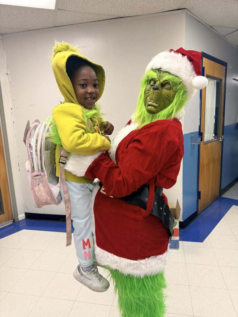 A person in a Grinch costume holds a smiling student while standing in a school hallway.
