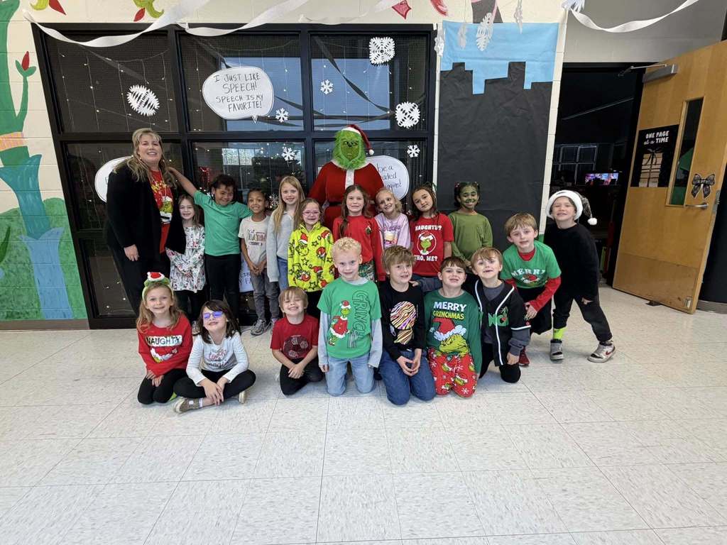 Students wearing Grinch-themed outfits stand and sit in a school hallway and pose with a teacher and an adult wearing a grinch costume.