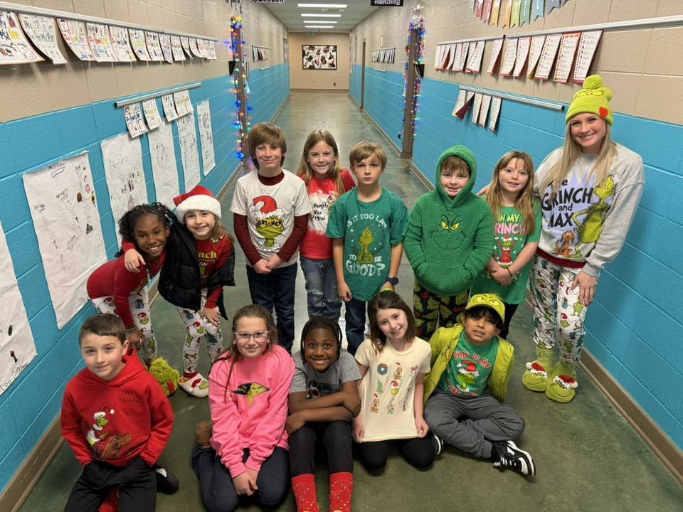 Students and a teacher dressed in Grinch-themed outfits smile for a photo while standing and sitting in a school hallway.