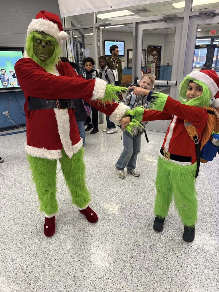 A child and an adult, both dressed like the Grinch point at eachother while standing inside a school hallway.