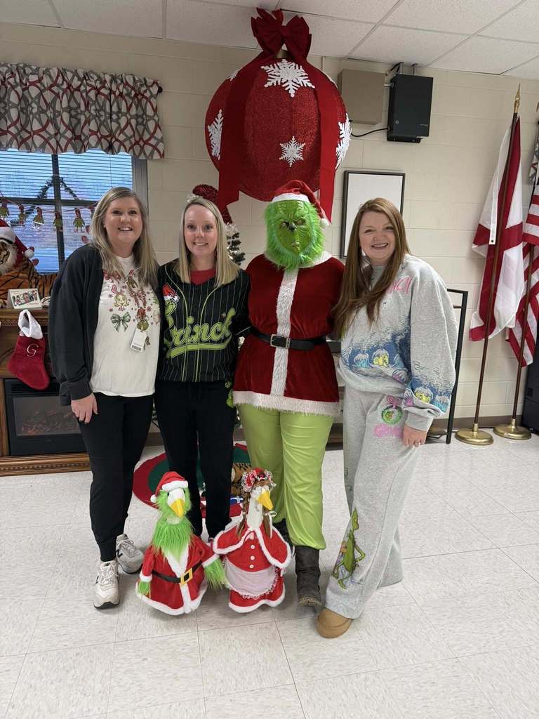 Teachers stand in a school and smile while posing next to someone in a Grinch costume. In front of the people, two ceramic geese sit on the floor wearing Christmas-themed outfits.