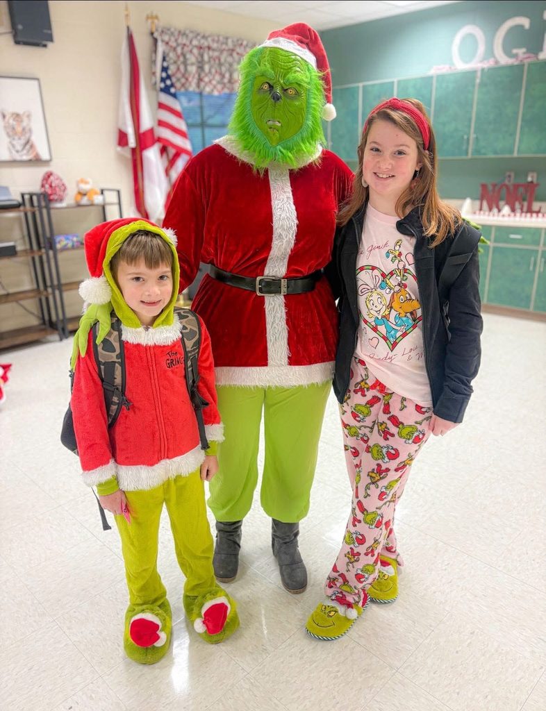 Children wearing Grinch-themed outfits pose next to someone in a Grinch costume while standing inside a school.