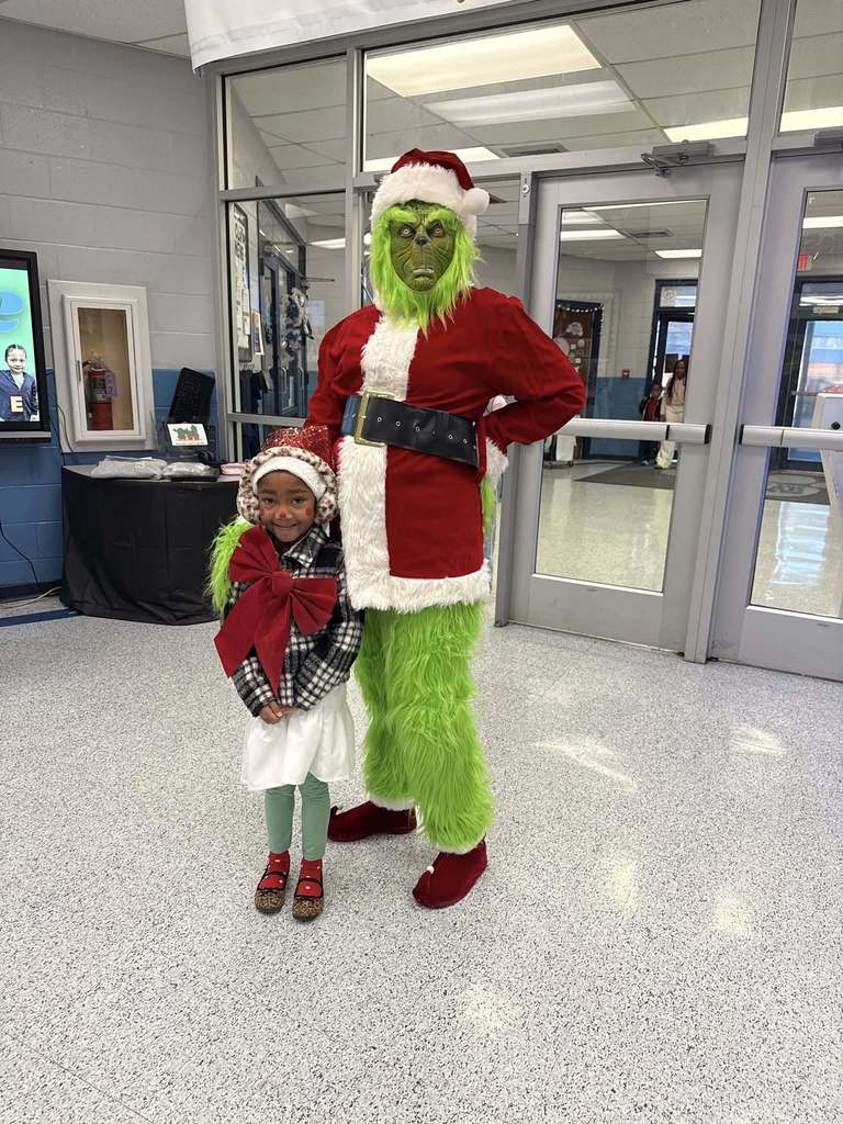 A student wearing a holiday outfit stands in a school hallway and poses next to an adult wearing a Grinch costume.