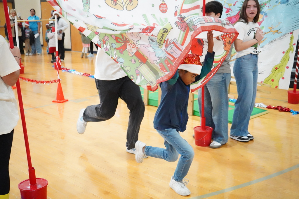 An elementary student and a high schooler run through a banner during an obstacle course set up in a school gym.