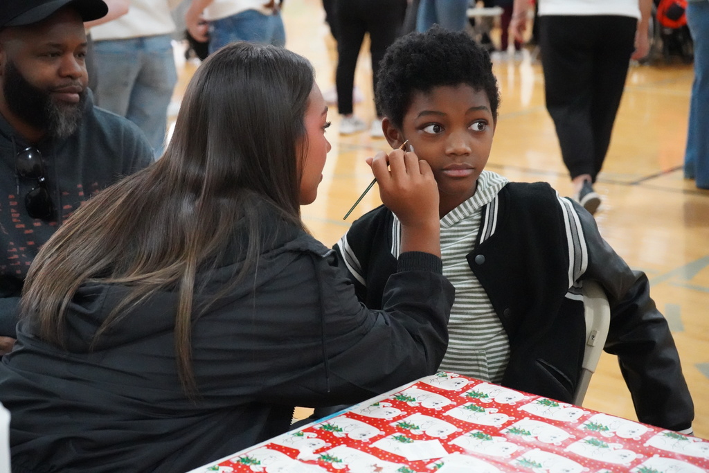 A high school student does face paint on an elementary school student while inside a school gym.