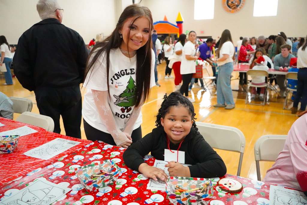 An elementary student and a high school student smile for a photo while doing arts and crafts inside a school gym.