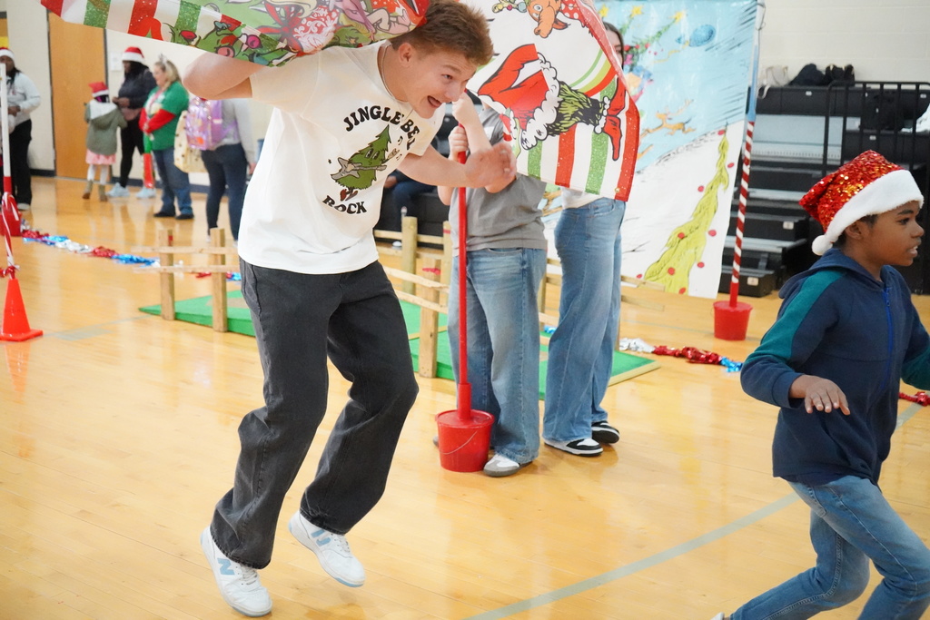 An elementary student and a high schooler run through a banner during an obstacle course set up in a school gym.