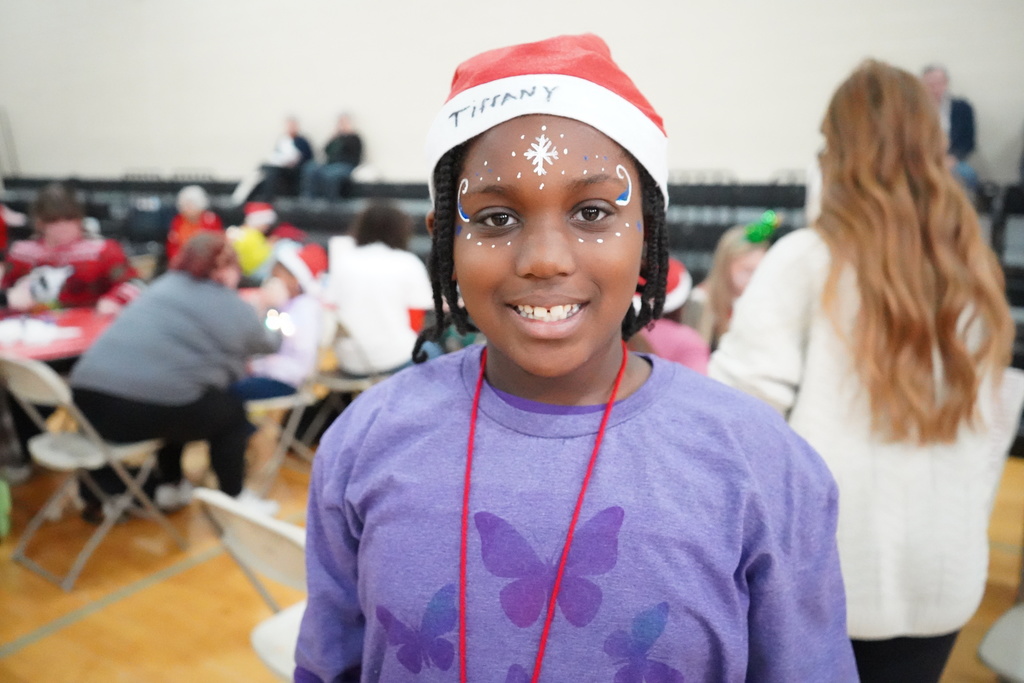 A student wearing a santa hat and face paint smiles for a photo while standing inside a school gym.