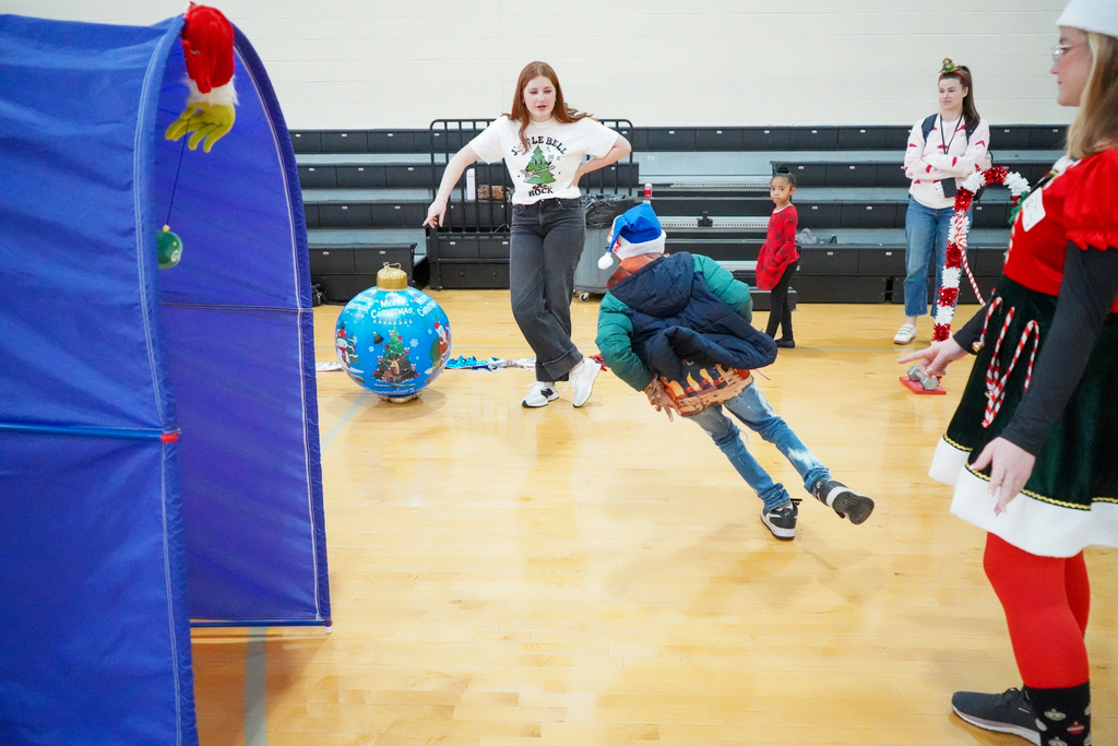 A student wearing a santa hat runs through an obstacle course set up inside a school gym.
