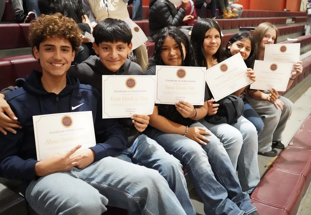 Six students smile for a photo following an award ceremony inside a school gym. All of the students sit on bleachers and hold up certificates.