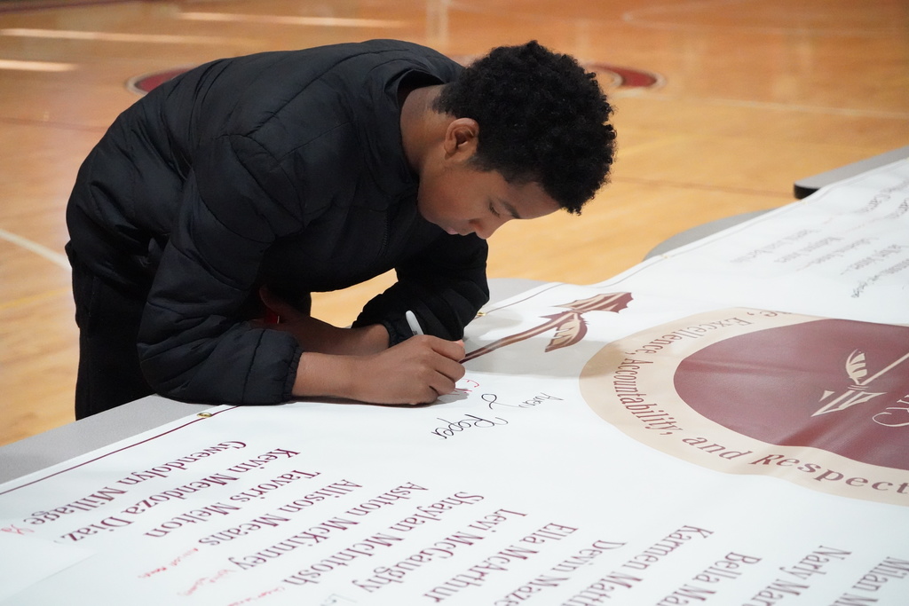 A student signs a banner during an award ceremony inside a school gym.