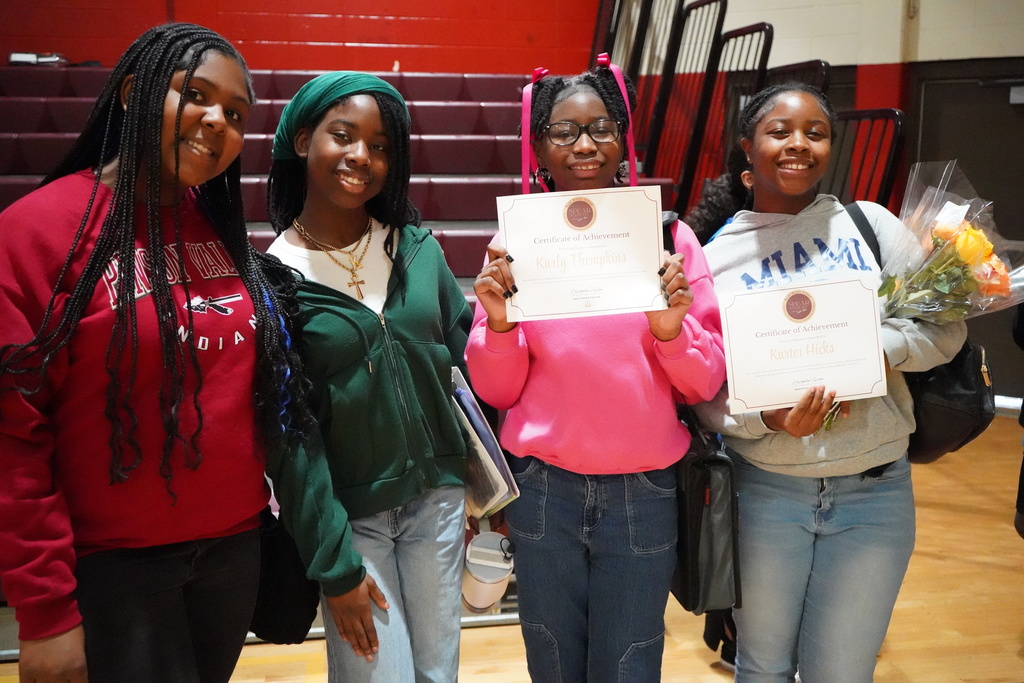 Four students smile for a photo following an award ceremony held in the school gym. One student holds a certificate and another student holds a certificate and flowers.
