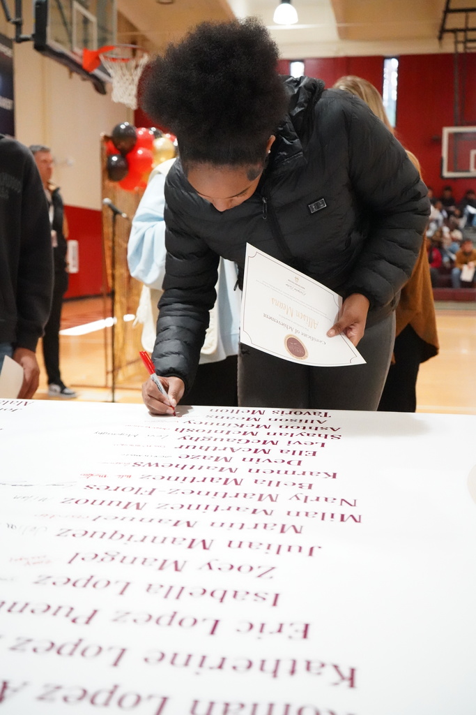 A student signs a banner during an award ceremony inside a school gym.