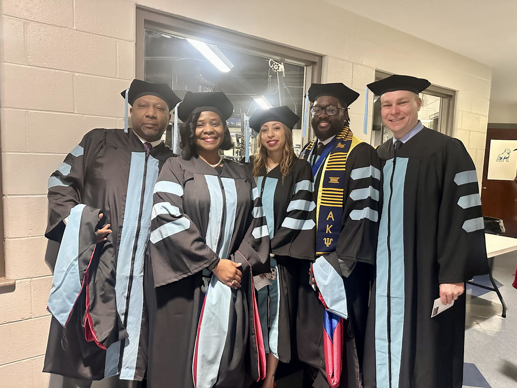 Dr. Roderigo Oliver, Dr. Nasiha Gresham, Dr. Suhai Douglas, Dr. Courtney Davis, and Dr. Hunter Davis stand together for a photo while wearing blue and black graduation caps and gowns. inside a building.