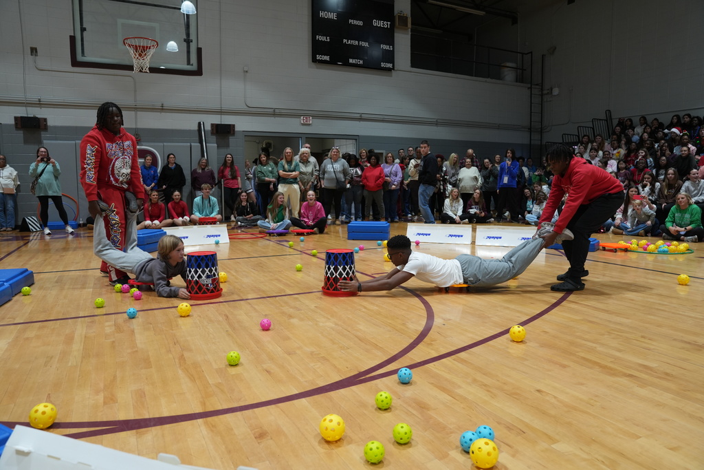 Image is from the Bragg Middle School Christmas Visions Pep Rally inside Bragg's gym. In the photo, students are playing a game where they try to put balls in a trash can while someone holds their legs.
