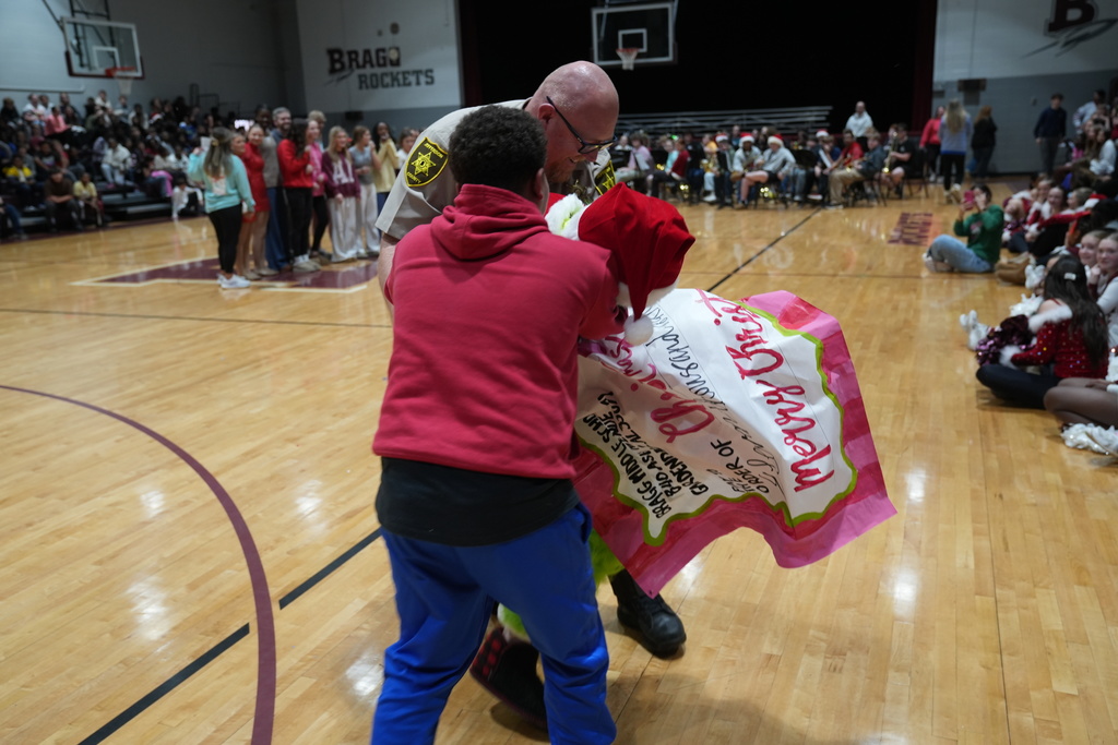 Image is from the Bragg Middle School Christmas Visions Pep Rally inside Bragg's gym. In the photo, a student and an SRO are stopping the Grinch from stealing a  donation.