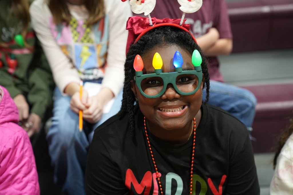 Image is from the Bragg Middle School Christmas Visions Pep Rally inside Bragg's gym. In the photo, a student wears Christmas themed glasses and smiles while sitting on the bleachers.
