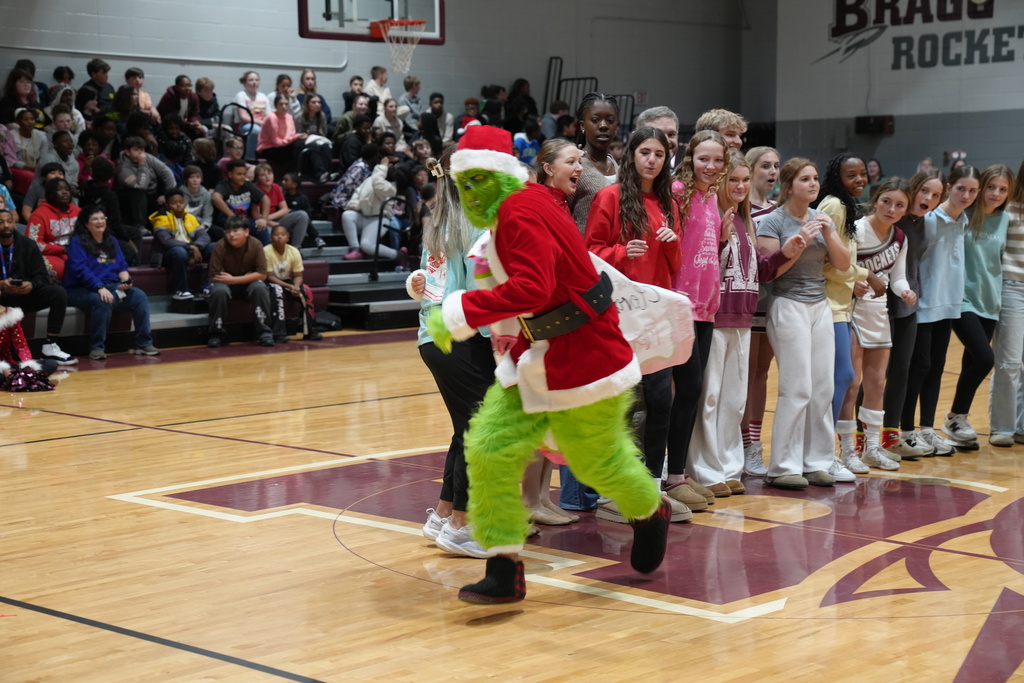 Image is from the Bragg Middle School Christmas Visions Pep Rally inside Bragg's gym. In the photo, the Grinch is stealing a paper with a donation amount written on it from students.
