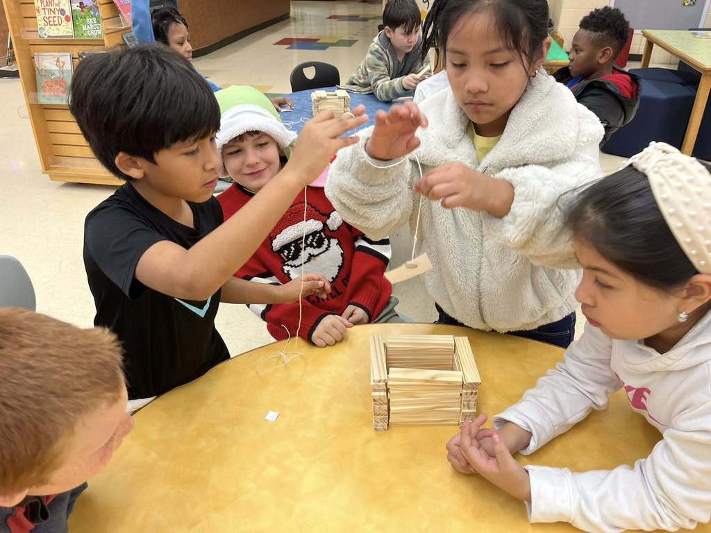 Irondale Community School students stand and sit at a table while two of the students dangle string with paperclips on it toward a structure made of blocks.