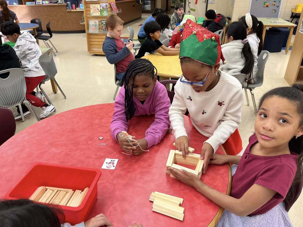 Three Irondale Community School students sit and stand at a table in the library. Two students work on a small structure made of wooden blocks. One of the students holds a string.