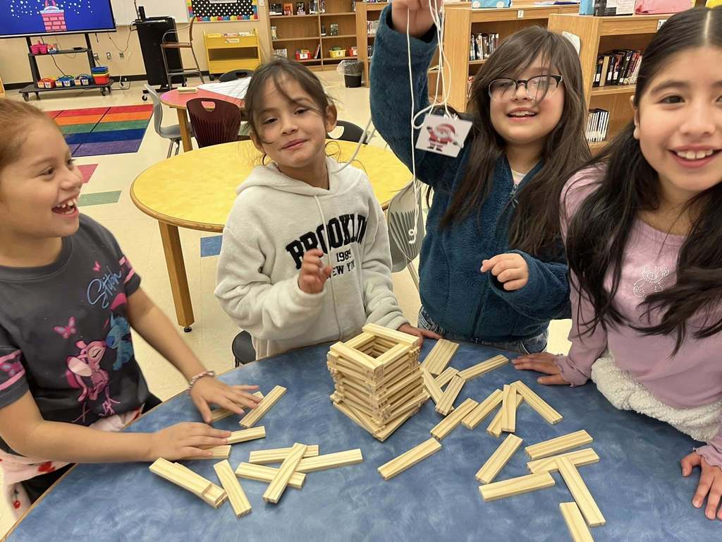 Students stand at a table with blocks on it and smile. One of the students holds a string with a picture of Santa on the end of it.