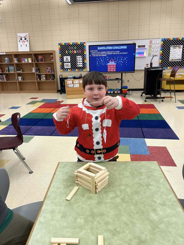 An Irondale Community School student holds a piece of string toward a structure made of blocks and smiles. The student is standing at a table in a library and is wearing a Santa costume.