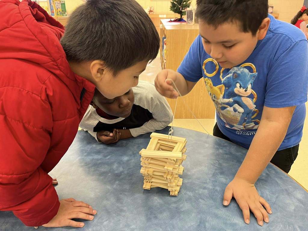 Three Irondale Community School students stand at a table and watch  as one of the students dangles a paper clip on a string toward blocks stacked in a tall square formation.