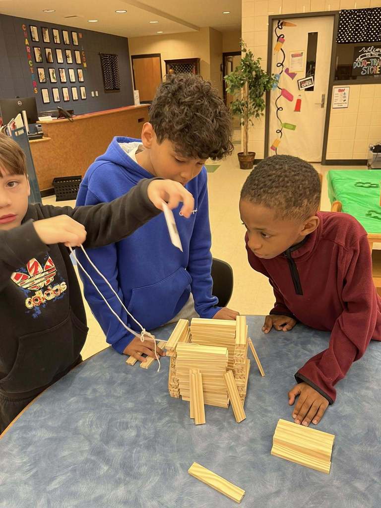 Irondale Community School students stand at a table in the library. Two of the students stare at a structure on the table made of wooden blocks. Another student holds up a string and a piece of paper.
