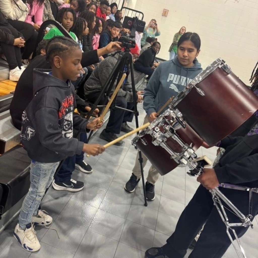 A young student plays a drum in a school gym while a band student holds it.