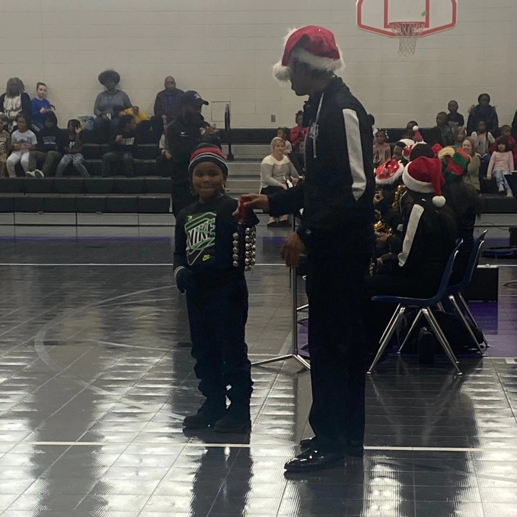 A band member holds out bells for a young student to see while they stand inside a school gym. 