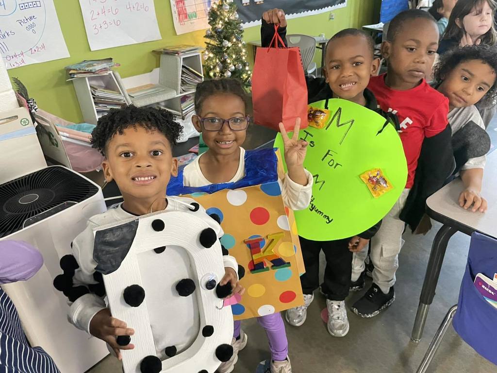 Students wearing costumes with different letters on them stand in a line in a classroom and smile.