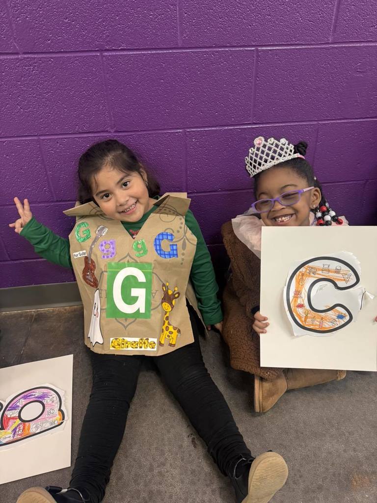 Students wearing costumes with letters on them sit in a hallway and smile.