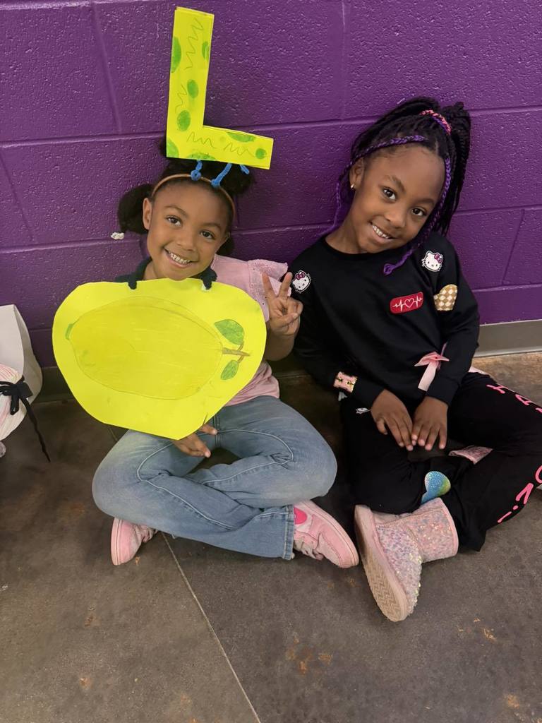 Students wearing costumes with letters and items representing that letter sit in a school hallway. One student has a yellow L on her head with a lemon drawing over her clothes. Another student has the letter H, Hello Kitty and Hearts on her clothes.