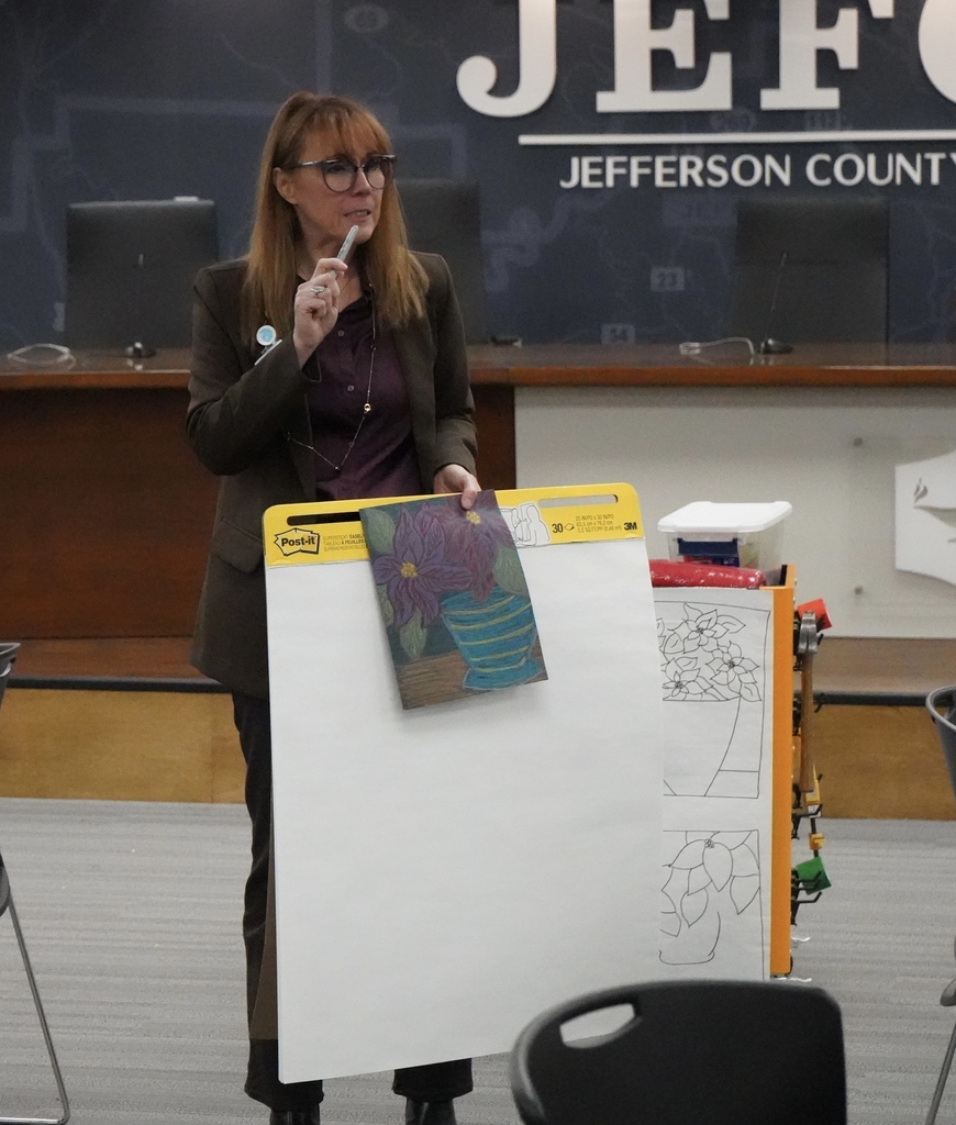 Arts Supervisor Shelly Bailey leads an art demonstration at the Jefferson County Schools Board of Education. She stands in the board room holding a drawing of poinsettias and a large notebook.