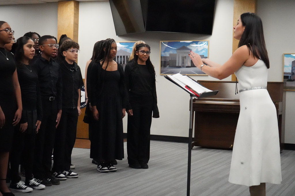 JCIB Middle Years Programme choir performs in the Jefferson County Schools Board of Education board room. The students are standing on the floor, singing while their choir director, Ms. Diana Preoteasa, conducts.