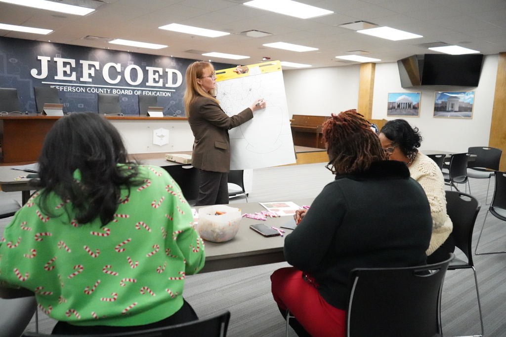 Arts Supervisor Shelly Bailey draws on a large notebook while leading an art demonstration in the Jefferson County Schools board room. Three women sit at a table in front of her drawing on construction paper.
