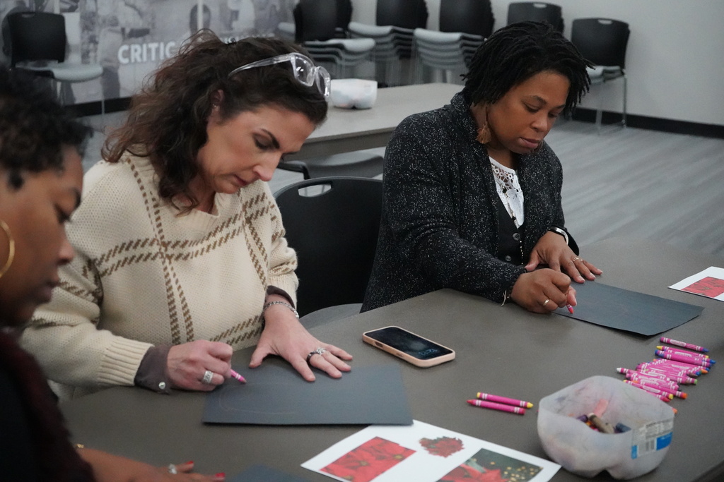 Three women sit at a table and draw on construction paper with crayons.