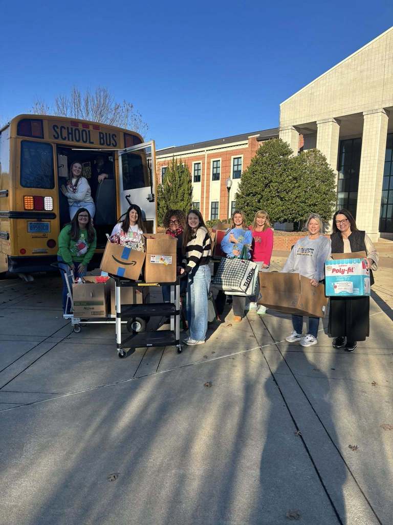 A group of women stand together and smile while loading boxes into a school bus.