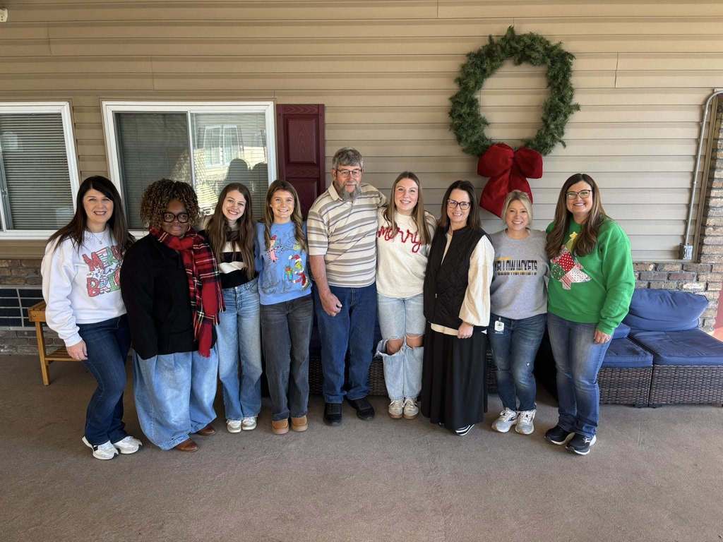 A group of women and a man stand together for a photo outside a building with a wreath on the wall.