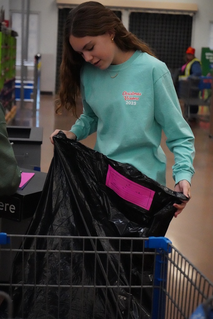 A student wearing a Christmas Visions shirt holds a garbage bag while standing inside a Walmart.