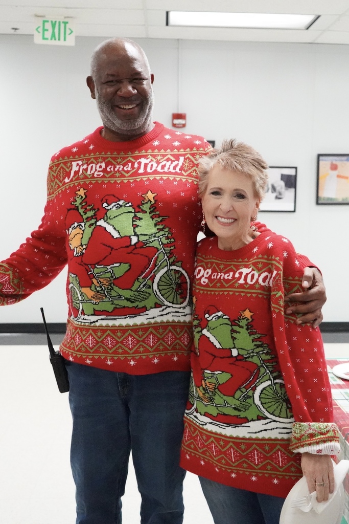 A man and a woman stand together for a photo and smile. They wear matching Frog and Toad Holiday Sweaters.