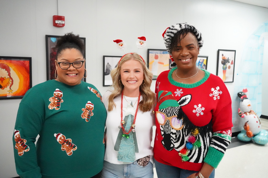 Three women stand together for a photo and smile. They all wear holiday sweaters. 