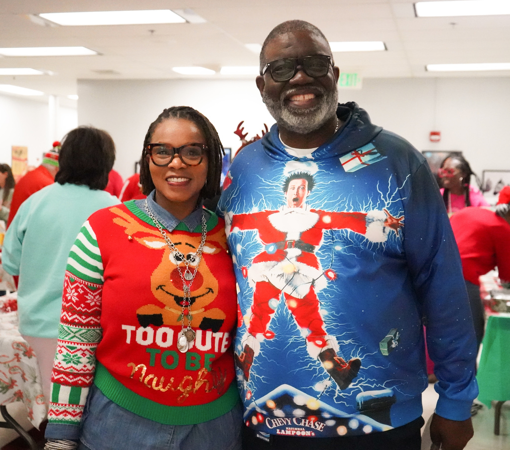 A man and a woman wearing holiday sweaters stand together for a photo and smile.